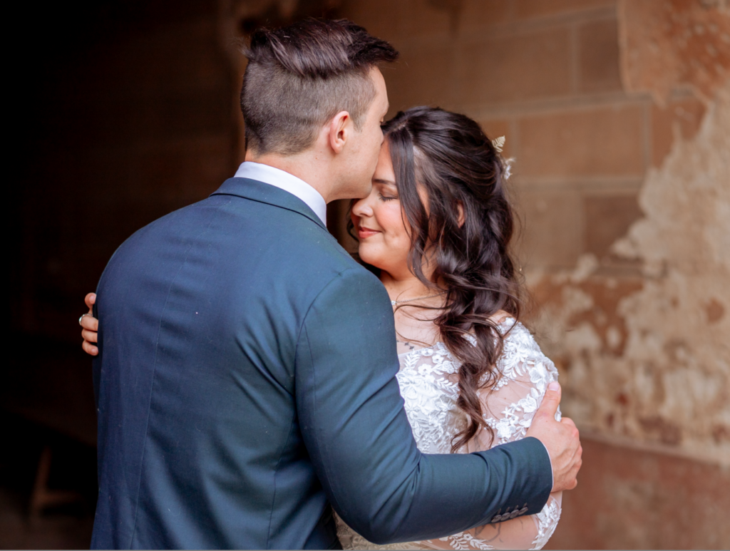 Photo de couple de mariés prise par May Jolies Photo, photographe de mariage. Le marié de dos embrasse la mariée, de face, sur le front. Photo douce, tons chauds et naturels