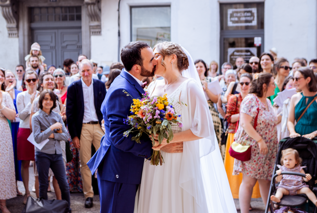 Photo d'un couple de mariés à la sortie de l'église, prise par May Jolies Photo, photographe de mariage. Le couple s'embrasse sur le perron de l'église devant tous leurs invités qu'on voit de face. Photo à l'ambiance joyeuse